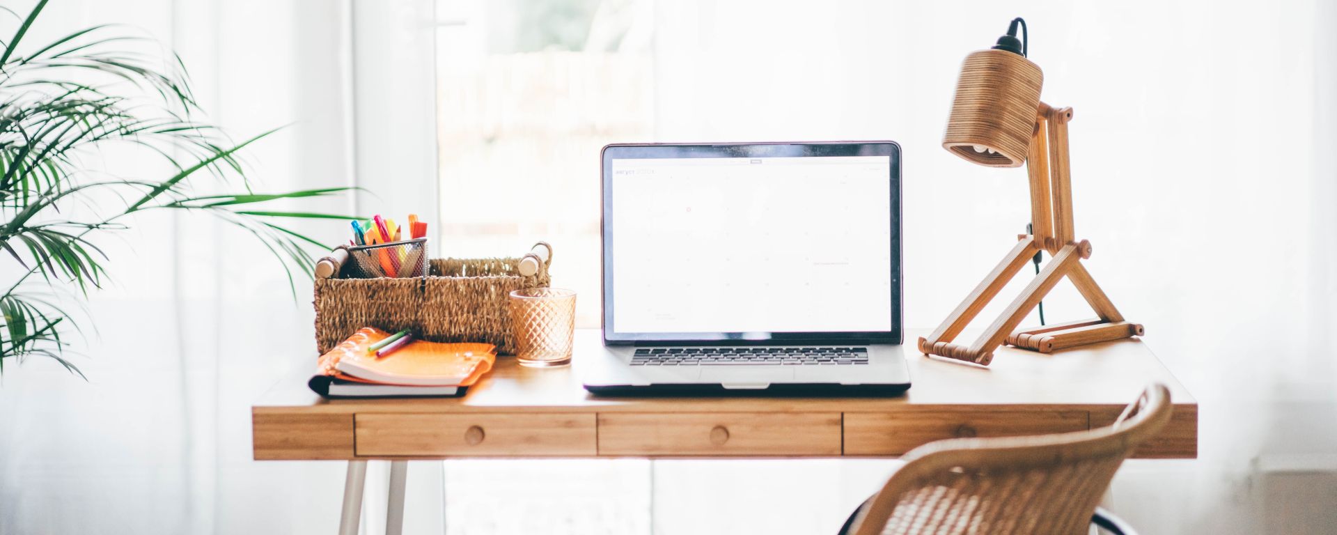 Organised home office with laptop, lamp, and stationary on a desk with good natural lighting in the background.