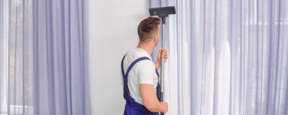 A man with a white shirt and blue overall vacuuming curtains